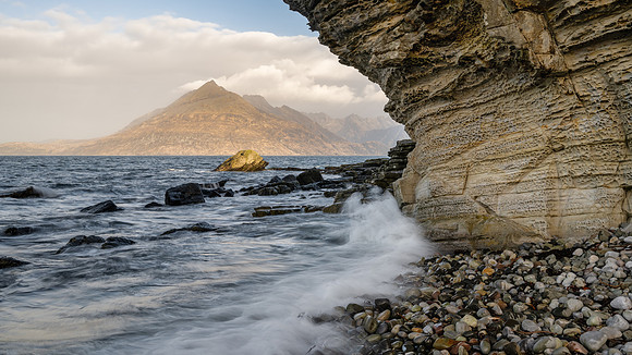 Waves crash on a pebble beach at Elgol with the Cuillins lit uo by the morning sun on the horizon