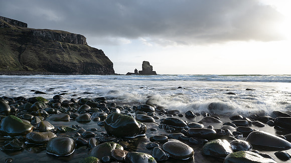 Low sunset light shimmers off the shiny black pebbles and basalt columns standing high on the cliff tops above Talisker Bay on the Isle of Skye