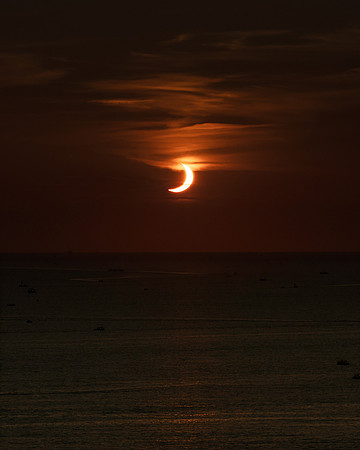 Observers slowly begin to depart in the boats as the setting sun emerges from behind the moon.