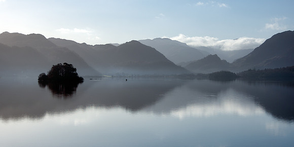 A man fishing on Derwentwater lake in a canoe at sunrise with mist on the water.