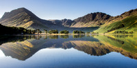 Mountains and trees reflected in still water at Buttermere, Cumbria during sunrise.
