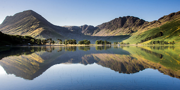 Mountains and trees reflected in still water at Buttermere, Cumbria during sunrise.