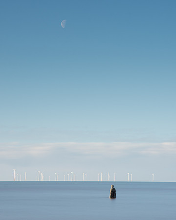 Statue of a man standing in sea looking at the moon over wind turbines at Another Place by Anthony Gormley.