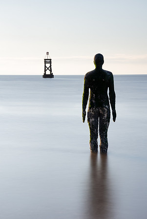 Statue of a man stands in the sea looking at the pier marker at Anthony Gormleys Another Place near Liverpool.