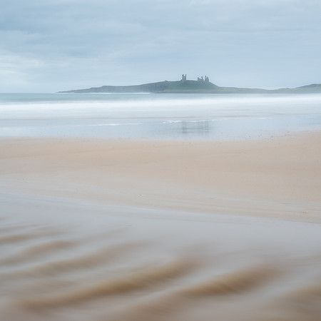 Ripples in the sand from the stream across Embleton bay beach across the waves with Dunstanburgh castle in the background.