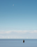 Statue of a man standing in sea looking at the moon over wind turbines at Another Place by Anthony Gormley.