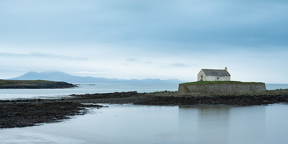 Welsh Mountains and Porth Cwyfan Church reflected on the sea at high tide in Wales.
