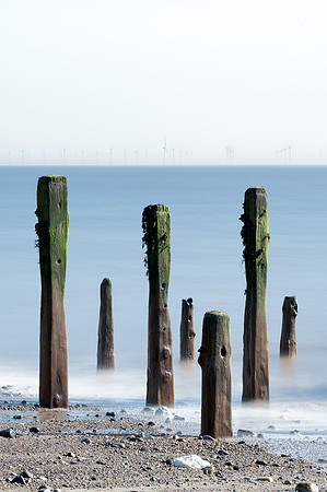 Tall posts from an old derelict jetty stand in sea at the shore with wind turbines on the horizon.