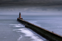 Long sea wall leads out to Tynemouth lighthouse under dark skies.