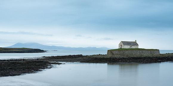 Welsh Mountains and Porth Cwyfan Church reflected on the sea at high tide in Wales.