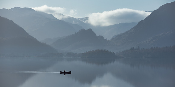 A man sails across Derwentwater lake in a canoe at sunrise with mist on the water.